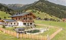 A beautiful wooden house surrounded by green meadows and mountains. In the foreground, there is a pool, surrounded by a wooden fence.