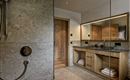 A modern bathroom with an elegant shower and a wooden vanity. The walls are clad in gray tiles and there is a large mirror light.