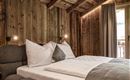 A cozy bedroom with wooden walls and a modern bed.  
Large windows let in daylight and provide access to a balcony.
