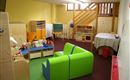 A bright playroom with green and blue furniture. There are toys, a table, and a staircase in the background.
