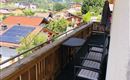 Ein Balkon mit Blick auf grüne Berge und ein kleines Dorf. Es gibt Stühle und einen Tisch auf dem Balkon.