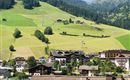 Eine malerische Landschaft mit grünen Wiesen und Bergen im Hintergrund. Im Vordergrund sind zahlreiche Dächer von Häusern und einige Tennisplätze sichtbar.