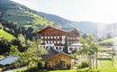 A picturesque hotel in the mountains, surrounded by green meadows and trees. The sun is shining, creating an inviting atmosphere.