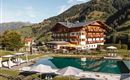 A picturesque hotel in the mountains with a pool and sun loungers in the foreground. Surrounded by green meadows and beautiful landscapes.