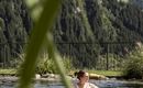 A person relaxes in a clear pool in front of a picturesque mountain landscape. The surroundings are green and sunny.