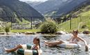 Two people are relaxing in a pool with a view of the mountains. The sky is clear, and the landscape is green and hilly.