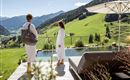 A relaxing spa atmosphere with a view of green mountains and a tranquil landscape. Two people in bathrobes enjoy the view next to a pool.