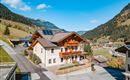 A charming house in the mountains with colorful flowers on the balcony. Surrounded by green meadows and impressive mountains under a blue sky.