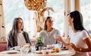 Three women are sitting at the table and enjoying breakfast together. In the background, there are windows overlooking a green landscape.