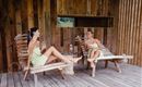 Two women are sitting relaxed on wooden loungers and having a conversation. The background is made of wood and conveys a cozy, natural atmosphere.