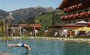 A man jumps into a clear, blue pool in the mountains. In the background, there is a building with flower balconies and a green landscape.