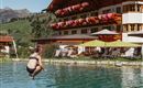 Eine Person springt in einen Pool vor einem hübschen Hotel mit blühenden Balkonpflanzen. Im Hintergrund sind grüne Berge zu sehen.