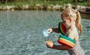 Ein Mädchen spielt im Wasser und gießt mit einem kleinen Behälter. Sie trägt bunte Schwimmflügel und hat blondes Haar.