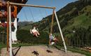 Zwei Kinder schaukeln fröhlich auf einer Spielplatzschaukel in einer malerischen Landschaft. Im Hintergrund sind grüne Hügel und ein bewaldeter Hang zu sehen.