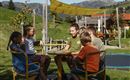 Eine Gruppe von Kindern sitzt auf einem Spielplatz und spielt an einem Karussell. Im Hintergrund sind die Berge und eine grüne Wiese zu sehen.