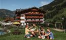 Vier Kinder spielen auf einer Wiese und halten Kaninchen in der Hand. Im Hintergrund steht ein wunderschönes Haus mit Balkonblumen und einer malerischen Berglandschaft.