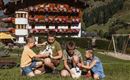 Vier Kinder sitzen auf der Wiese und spielen mit Zwergkaninchen. Im Hintergrund ist ein schön dekoriertes Gebäude mit Blumen vor einer grünen Berglandschaft zu sehen.