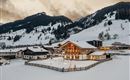 A picturesque mountain village in winter, surrounded by snow-covered hills. Cozy houses with warm light stand out against the cloudy sky.