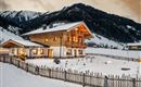 A charming wooden house in winter with a snow-covered garden. In the background, the mountains and a cloudy sky can be seen.