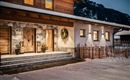A cozy wooden house in the snow with an inviting front door. The facade is modern and stylish, surrounded by mountains and a tranquil winter landscape.