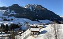 Eine Winterlandschaft mit schneebedeckten Hügeln und Bergen im Hintergrund. Kleine Häuser liegen in einem ruhigen Tal unter klarem blauen Himmel.