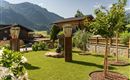 A picturesque garden with green lawn and well-maintained trees. In the background, mountains and traditional houses are visible.