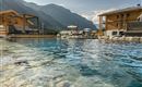 A pool with clear water, surrounded by pretty wooden cabins in a mountainous landscape. In the background, the gentle mountains and a blue sky are visible.