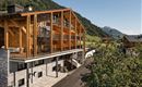 A modern hotel building made of wood and stone in a picturesque mountain landscape. The surroundings are green and inviting, with views of gentle hills.